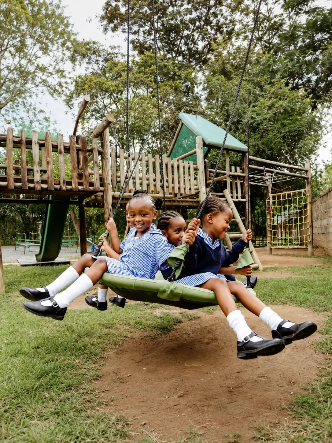 Three JENS learners in school uniform laughing together on a large green swing on the school playground